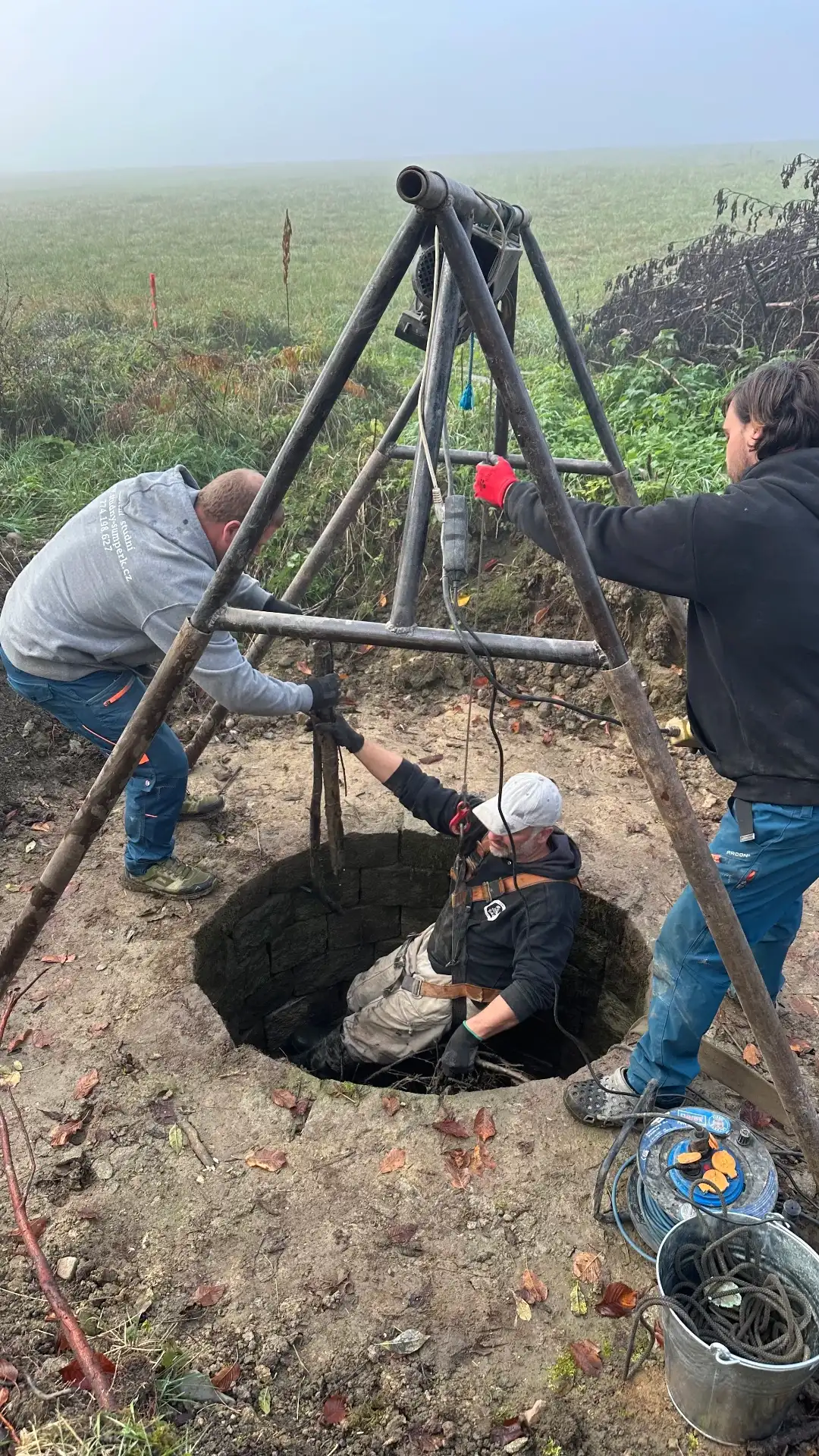 Cleaning the historic well — three people with a tripod, misty morning in the Rychleby Mountains